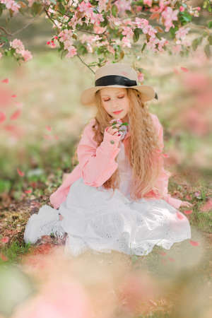 Nice longhaired blonde girl in white dress, pink jacket and straw hat set under the tree in blooming apple garden. Vertical photo.の写真素材