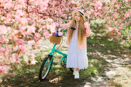 Nice longhaired blonde girl in pink jacket and straw hat walking in blooming apple garden with retro bicycleの写真素材