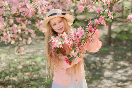 Nice longhaired blonde girl in pink jacket and straw hat hold blooming branch of apple tree covered by flowers. Horizontal photo.の写真素材