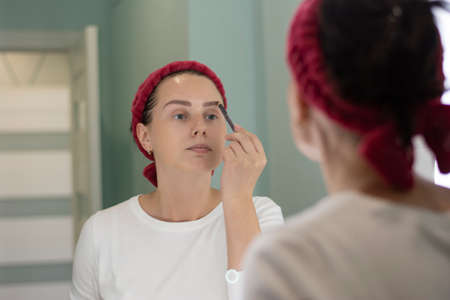 Young beautiful brunette girl dyes her eyebrows at home with hair bandages looking in a mirror reflectionの写真素材
