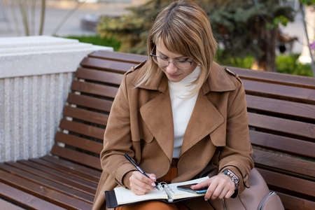 Beautiful blonde businesswoman in glasses sitting on a city street bench writes notes in a notebookの写真素材