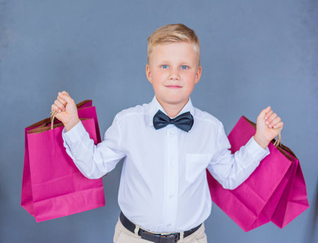 Close-up of a blond boy in a white shirt with a bow tie holds two gift bags on a blue background isolateの写真素材
