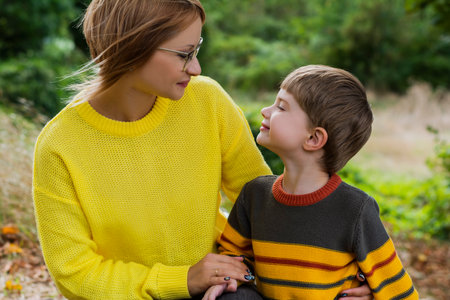 Mother and son in a cozy yellow brown sweater are sitting in the forest in autumn on a sunny day smiling looking straight into the camera picnic rest on nature weekendの写真素材