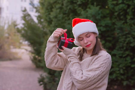 Close up woman in Christmas cap shaking red box at her ear trying to guess what is inside standingの写真素材