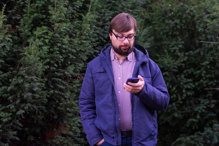 Man with glasses and a beard is typing a message on his smartphone on the street near the treesの写真素材