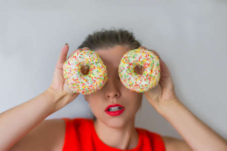 Joyful beauty model woman with red lipstick and red dress holding donuts near eyes on backgroundの写真素材