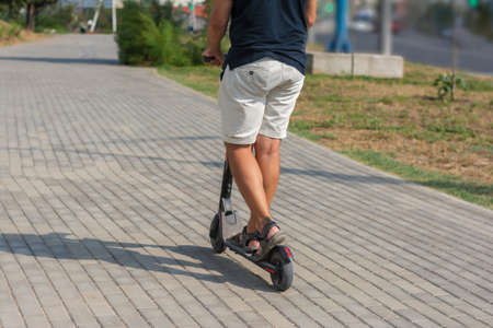 Close-up legs of a man on a electric kick scooter in a park on the road in summer at sunset