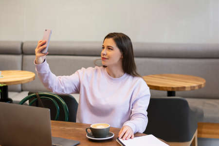 Beautiful young woman holding her mobile phone and taking selfie or videocalling in a cafe mock upの写真素材