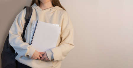 Faceless student in a beige sweatshirt with a backpack holds notebooks in his hands against a lightの写真素材