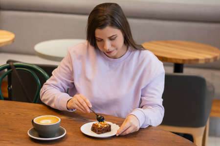 Enjoying morning routine beautiful millennial woman eating delicious brownie and drinking coffeeの写真素材