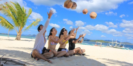 Two happy young couples sit on tropical white beach and play with coconuts as balls, with background of beautiful blue sky and cloud and ocean. Shot on Pass Island, Coron, Palawan, Philippinesの写真素材