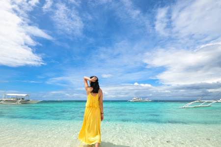 Back portrait of single young woman in yellow long dress walks in shallow water on tropical beach の写真素材