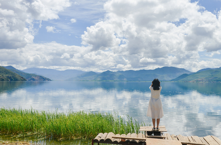 Single young Chinese woman in white long skirt standing near the Lugu lake, listening to music, rear view, at Daluoshui village, Lijiang, Chinaのeditorial素材