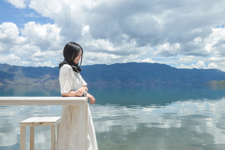 Single Chinese young girl looks aside, smiling, leaning on white long table by the bank of Lugu lake, at Daluoshui village, Lijiang, Chinaの写真素材