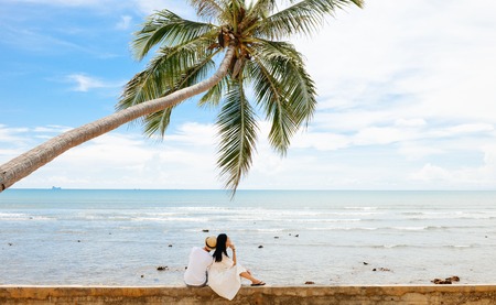 young Asian couple leans on each other under one palm tree at tropical beach, Lanta island, Krabi, Thailand.の写真素材