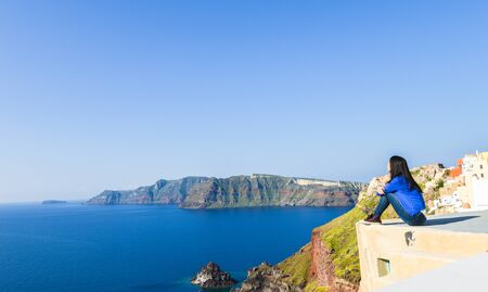 Single Asian girl sitting on edge of roof top and looking at horizon at Oia town, Santorini, Greeceの写真素材