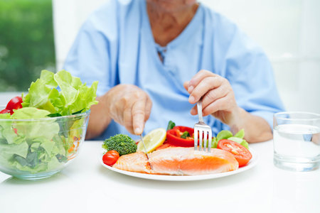 Asian elderly woman patient eating salmon stake and vegetable salad for healthy food in hospital.の写真素材