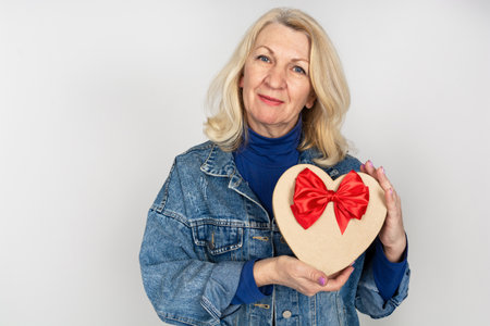 Beautiful elderly woman smiling and holding a heart-shaped gift box in her hand on a white background.の写真素材