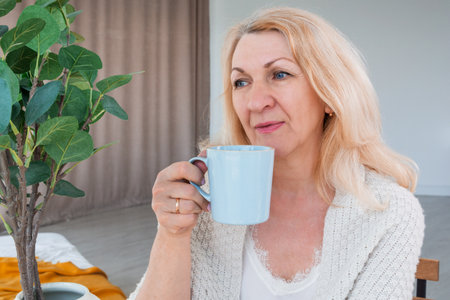 A pensive middle-aged blonde holds a cup of tea or coffee in her hand.の写真素材