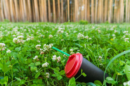 A black paper coffee cup with a straw is thrown into the green grass. Environmental pollution with household waste. Inscription in Russian: careful, hot.の写真素材