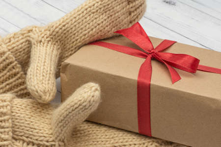 Knitted mittens hold a holiday gift on a white wooden background. The concept of congratulations to...