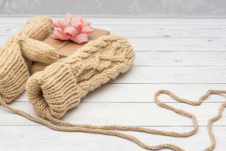 Knitted mittens hold a holiday gift on a white wooden background. The concept of congratulations to...
