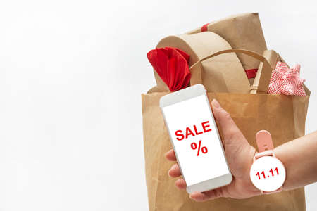 A woman with a wristwatch holds a smartphone in her hand against the backdrop of a large bag of gifts on a white background. Phone and clock have room for text.の写真素材