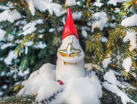A toy gnome in a red hat stands in the snow next to a tree and holds a star. Christmas and New Year concept.の写真素材