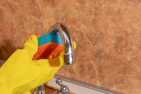 Woman in yellow rubber gloves washes a metal faucet with a sponge at the sink in the kitchen.