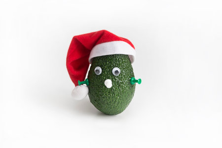 Avocado in a Christmas hat with a push pin instead of a nose on a white background. Funny avocado face. Christmas concept.の写真素材