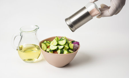 Prepared vegetable salad in a bowl on a white background. Nearby is olive oil in a glass jug. A woman's hand adds salt to the salad.の写真素材