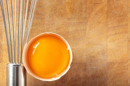 An egg yolk in its shell with a metal whisk on an old wooden chopping board.の写真素材