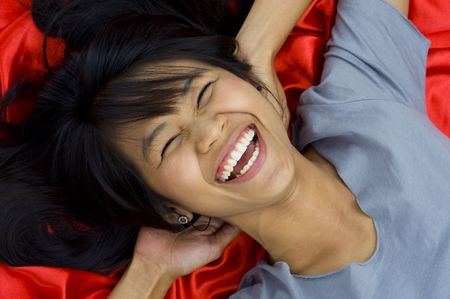 beautiful, young asian laughing with eyes close, laying on red shiny silkの写真素材