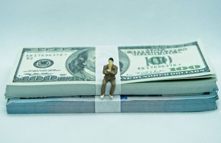 Figure of a man in a suit on top of a wad of hundred dollar bills in a white backgroundの写真素材
