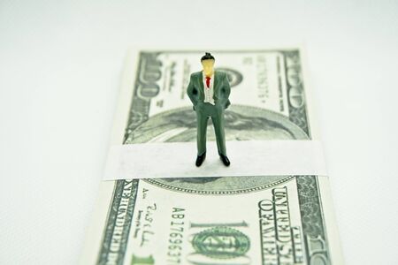 Figure of a man in a suit on top of a wad of hundred dollar bills in a white backgroundの写真素材