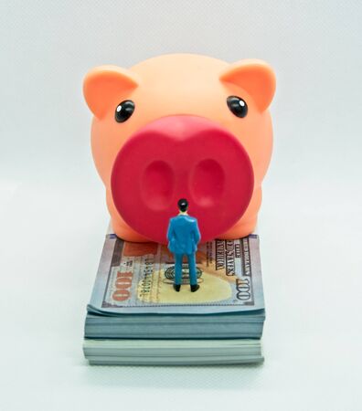 Piggy bank with a figure of a man in a suit in front of it and on top of a wad of hundred dollar bills in a white backgroundの写真素材