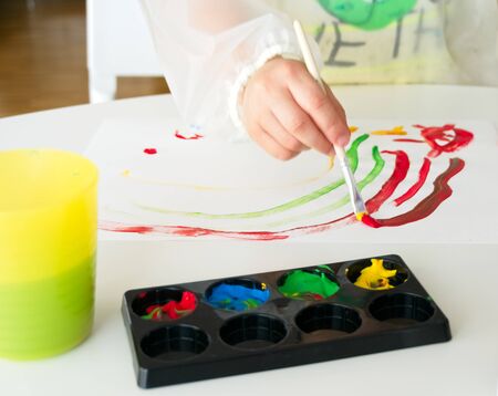Little boy painting with a brush and tempera paint a white paper on a white table with different containers with colored paintsの写真素材