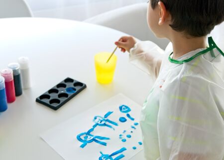 Little boy painting with a brush and blue paint and a white paper on a white table with different containers with colored paintsの写真素材
