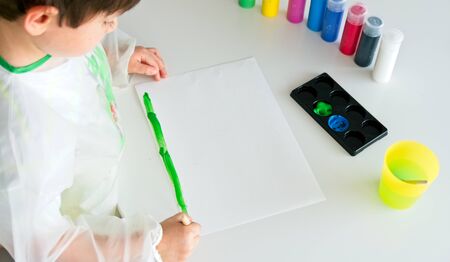 Little boy painting with a brush and a white paper on a white table with different containers with colored paintsの写真素材