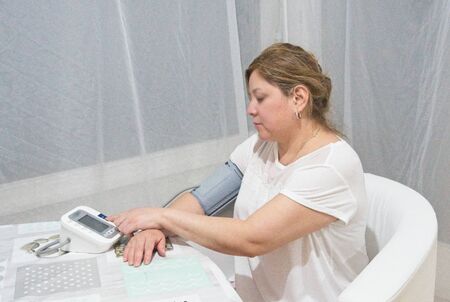 Woman sitting in a chair with her arm resting on a table while taking her blood pressure with an...