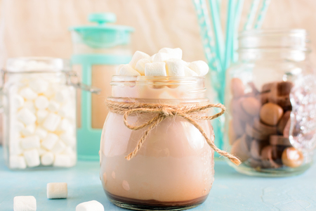 Cocoa in a jar, on top of a bunch of marshmallows. In the background, a blue coffee pot, chocolate sweets and turquoise cocktail tubes in a blur. Gentle still life.の写真素材