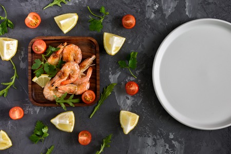 On a wooden plate are shrimp. Next on a black textured background, scattered herbs, spices and...