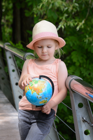A little girl in a straw hat is holding a globe in her hands. Child considers the model of the globe.の写真素材