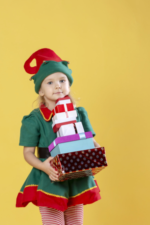 Little girl in a Christmas elf costume, carries a stack of gifts. Photo on a yellow background.の写真素材