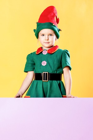 Little girl in a Christmas elf costume and in a red cap. Place for text on a pink background, copy...