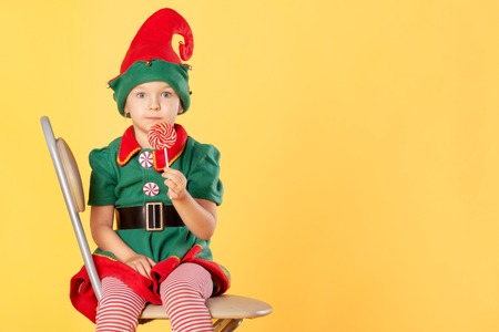 A beautiful baby in a suit of a Christmas elf is sitting on a chair. She has a delicious lollipop in her hands. Surprise. Near Copy Space.の写真素材