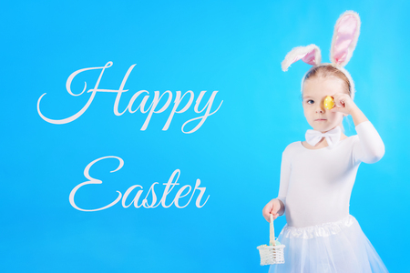 Child in a white rabbit costume. The little girl holds in one hand a basket and in the other hand an egg with which she covers the eye. Cute bunny, holiday symbol.の写真素材