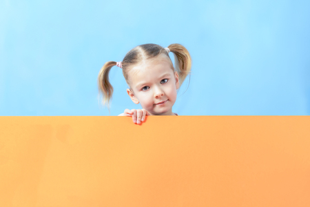 Copy space on a sheet of orange cardboard behind which stands a little girl. A cute child with blond hair. Photo with space for text and advertising.の写真素材