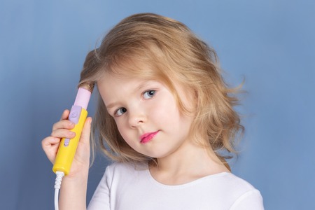 Little girl with blond hair makes styling. The child is holding curling irons.の写真素材