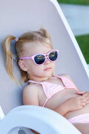 A little girl with blond hair sunbathes in sunglasses. The child is on vacation, lying on a sun lounger.の写真素材